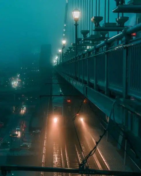 Three illuminated traffic lights hang on a bridge over a foggy city street at night, with a view of the illuminated downtown buildings in the distance