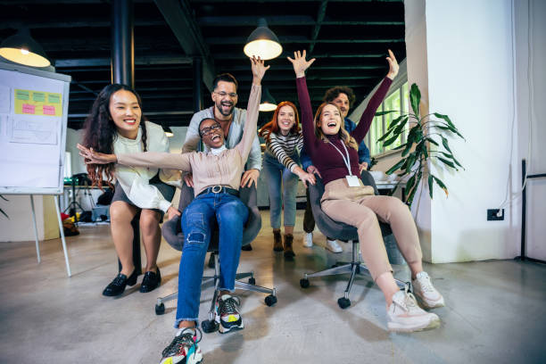 jovens empresários alegres em roupas casuais inteligentes se divertindo enquanto correm em cadeiras de escritório e sorrindo - office fun competition chair - fotografias e filmes do acervo