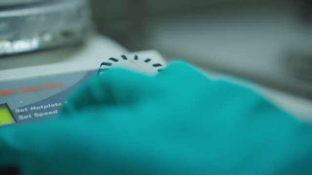 A scientist's hand in a protective glove adjusts stirrer speed and heating temperature of hot plate