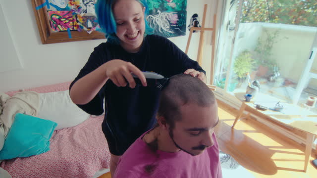 Happy Young Girl Shaving Head of Boyfriend with Electric Razor at Home