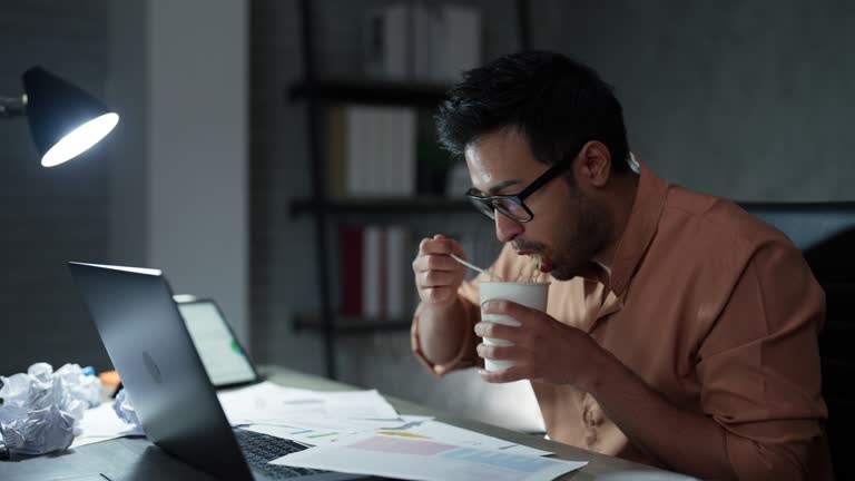 Asian young adult man is sitting at his desk to eat instant cup noodles while working on a project plan during overtime.