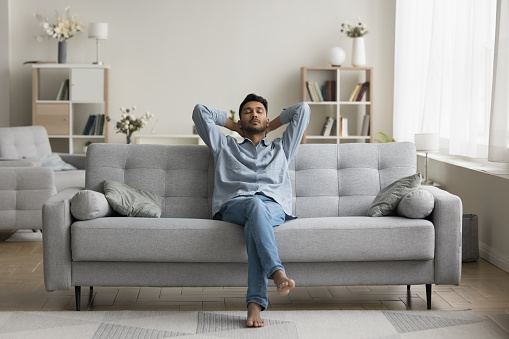 Serious calm young Indian man resting on home couch, sleeping, enjoying leisure time, silent peaceful break, pause, relaxing, sitting on comfortable sofa in modern cozy apartment