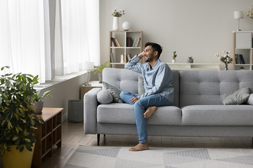 Positive dreamy relaxed young Indian guy sitting on comfortable couch, enjoying home leisure, relaxation, comfort, thinking, dreaming, planning, smiling, laughing, looking at window away