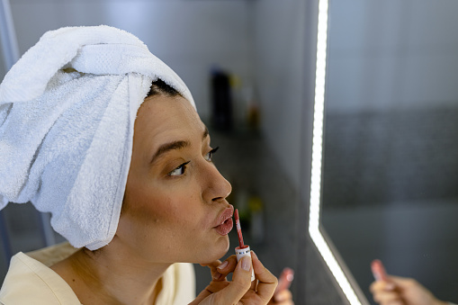 A beautiful young woman in a bathrobe is preparing for work and applying lipstick on her lips.