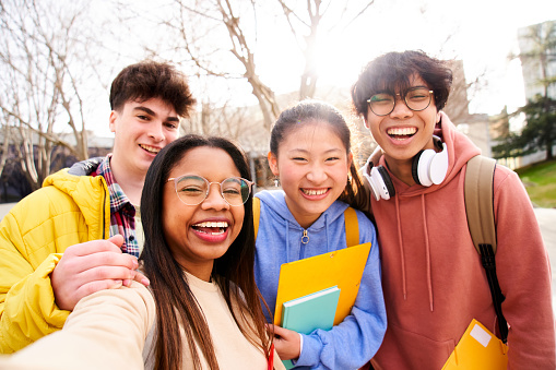 Group of multi-ethnic high school students taking a selfie outdoors at the university campus holding folders. Looking at camera picture of a diverse colleagues having fun.