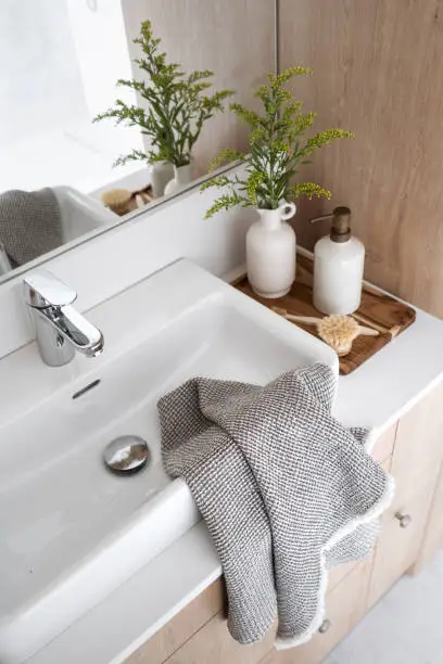 towel on porcelain washbasin, chrome water tap, massage brush, vase with plant and dispenser bottle with soap on wooden tray on countertop in domestic washroom, high angle shot