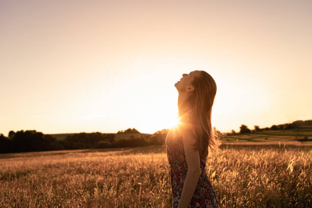 Gioiosa giovane bella donna in piedi in un campo al tramonto guardando il cielo sentendosi felice. - foto stock