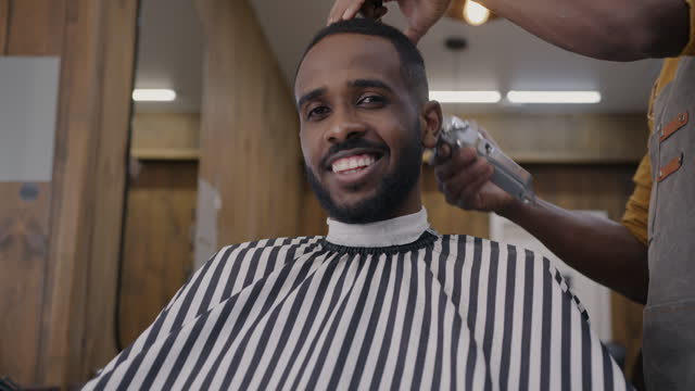 Portrait of cheerful African American man smiling looking at camera while barber shaving hair with electric machine