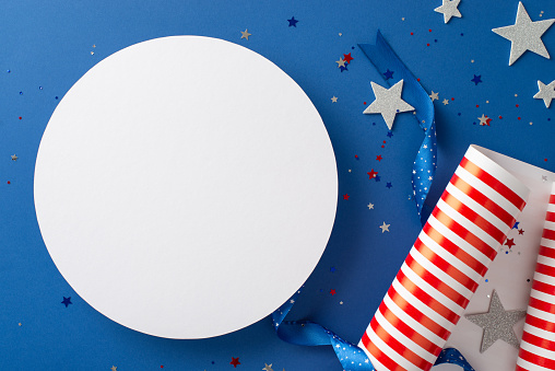 Celebrate Independence Day with grandeur. Top view shot of themed wrapping paper roll, ribbon, glittering stars and confetti on blue background, offering empty circle to include text or advertising