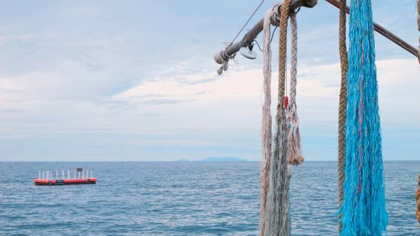 cordas e redes de pesca pendem sobre a superfície do mar, criando sensação de tranquilidade e harmonia com a natureza. pequeno barco de pesca que flutua longe da costa. as redes de pesca pendem de cima para baixo da viga. - fishing net commercial fishing net netting isolated - fotografias e filmes do acervo