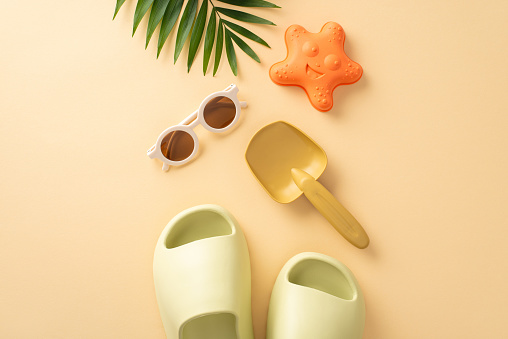 Tropical beach getaway with little one. Top view of beach toys, shovel, starfish sand mold, shades, slippers, palm leaf, soft beige backdrop. Perfect for promoting family-friendly vacation or product