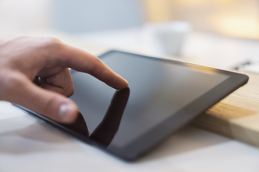 Close detail of a man's finger making contact with the screen of a sleek digital tablet resting on a desk, set against the soft-focus backdrop of a light-filled office