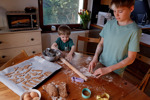 Children, boys, brothers with Vitiligo are sowing flour and kneading dough together at the table. Children laugh, play, have fun making cookies together. Flour is scattered on a wooden table, there are ingredients for dough and kitchen tools. Festive, homemade gingerbread cookies for the family.