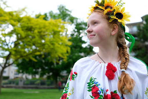 young red-haired girl in a wreath of sunflowers outdoors in park garden in beautiful embroidered Ukrainian vyshyvanka shirt with red braid with ribbons Ukraine Peaceful Time of happiness joy and peace