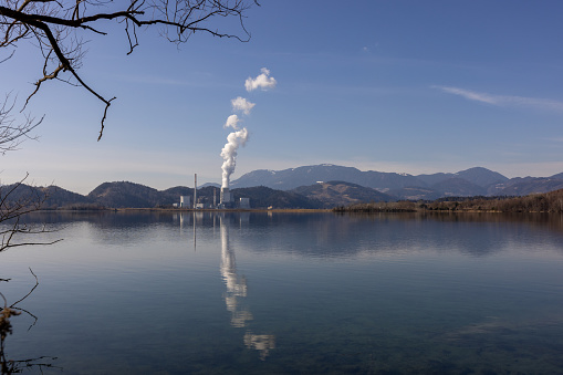 Smoke is rising from cooling tower of nuclear power plant by mountain with reflection on river water surface