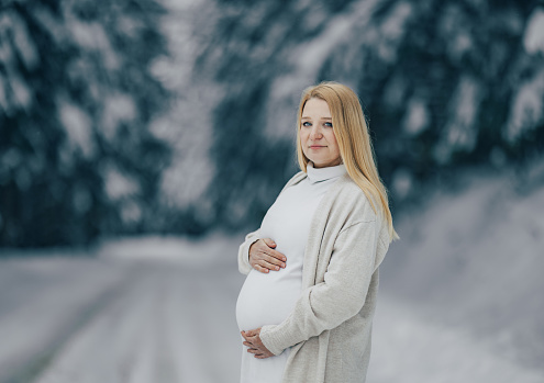 Beautiful woman touching her pregnant belly in warm clothing outdoors at snowy forest