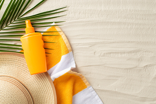 Sun protection on vacation concept. Top view photo of beach towel, straw hat, sunscreen spray and palm leaf on the sand on isolated background with copyspace