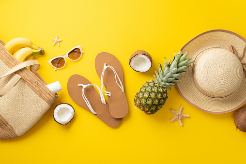 Captivated by the tropical paradise. Overhead view of beach bag, stylish eyewear, sunhat, sunscreen, flip-flops, starfish and succulent tropical fruits on a vivid yellow background