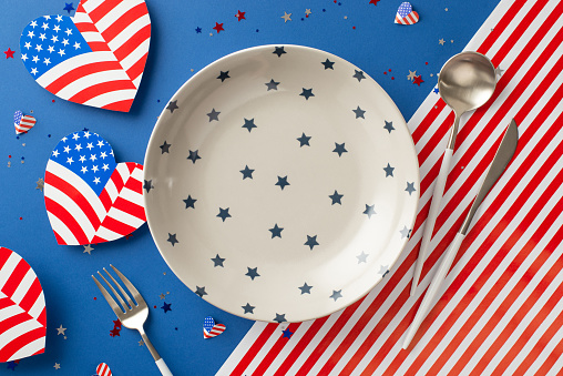 Organize menu for Fourth of July gathering. Top-view shot of festive table featuring empty plate, silverware, heart-shaped flags, and vibrant confetti on USA flag inspired backdrop with space for text