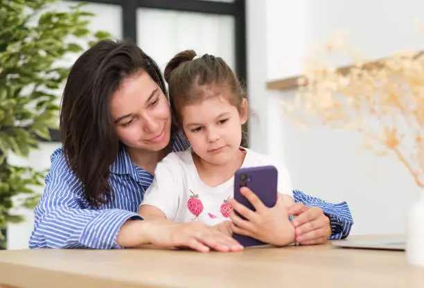 Photo of Head shot smiling young mother showing funny cartoons to overjoyed little adorable girl on smartphone. Happy 30s woman recording family video with laughing small preschool daughter on mobile phone.