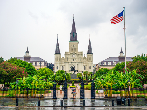 New Orleans, Louisiana, USA, August 27, 2017: A rainy day at the St. Louis Cathedral in New Orleans, Louisiana. A woman is seen about to cross the street holding a purple umbrella while a couple more people are seen in ponchos behind the gates.