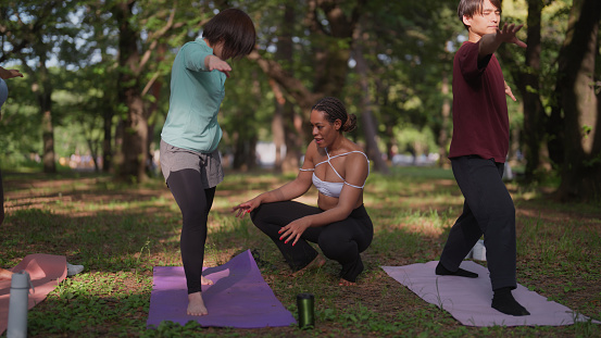 A group of multi-ethnic people are taking part in a yoga class in nature. A Black ethnic female yoga instructor is teaching yoga to a group of multi-ethnic people in nature. A Black ethnic female yoga instructor is fixing posture of yoga learners during yoga class in nature.