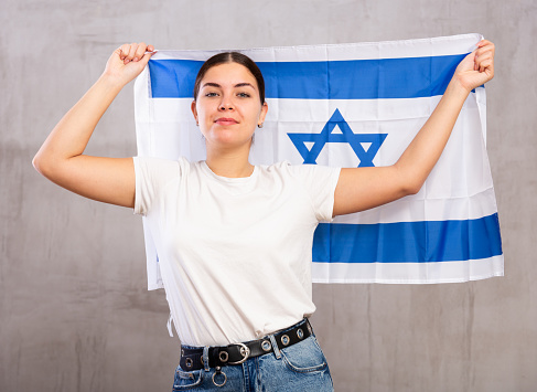 Portrait of serious patriotic young female holding national flag of Israel against gray wall background