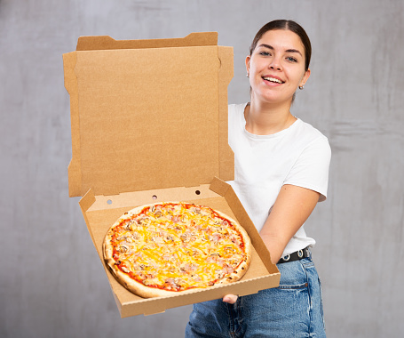 Photo of whole pizza held by cheery young woman against gray shadeless background