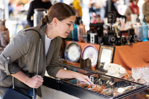 Young woman in casual clothes chooses vintage cutlery at flea market