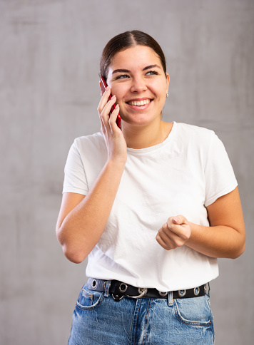 Photo of cheerful young woman speaking on mobile phone with pleasure against gray shadeless background