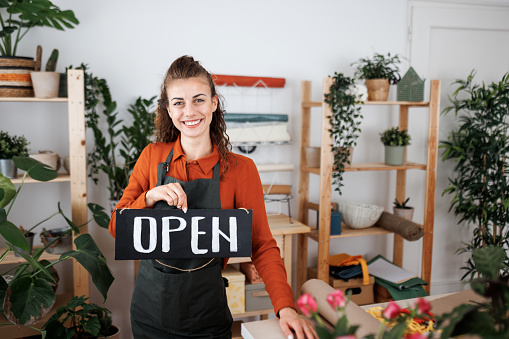 Young beautiful woman posing for a shot at her florist shop while holding open sign in her hand