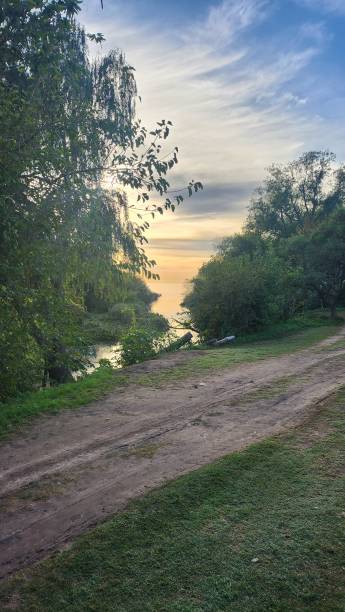 Diagonal dirt road in the foreground with trees at the river's edge, sunset with orange sky reflecting on the water, with clouds stock photo