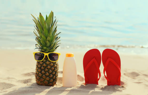 Summer vacation, stylish pineapple with sunglasses, red flip flops and bottle of suntan cream on the beach on sea background stock photo