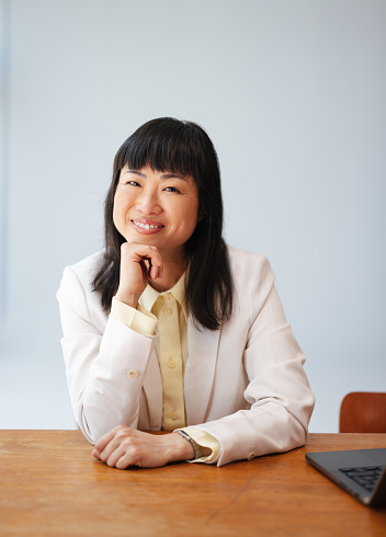 Close up shot of a beautiful Asian woman dressed in business wear, sitting at her desk at work. She is smiling while looking away. There is a laptop computer on the table and a notebook.