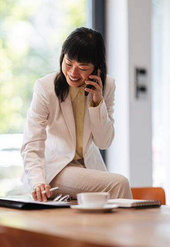 Happy Asian woman speaking on the phone with client and analyzing business report on a paper while sitting on her office desk with a laptop computer, notebook and cup of coffee.