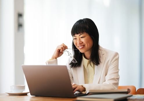 Smiling Asian woman analyzing business report on a laptop while sitting at her office desk with a cup of coffee and notebooks.
