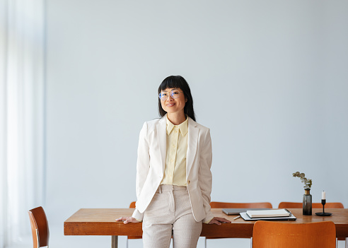 Beautiful Asian businesswoman, formally dressed, leaning against her office desk. She is wearing glasses, smiling and looking at the camera.