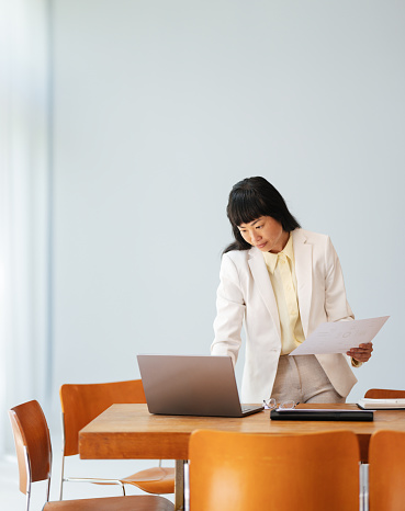 Beautiful Asian woman standing by her desk at work. She is holding a piece of paper and looking down at her laptop computer. She is working with contracts, reports, some research.