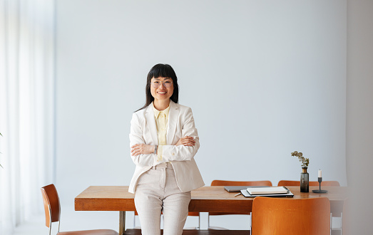 Smiling Asian business woman with crossed arms leaning on her office desk and looking at camera.