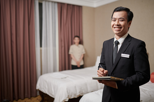 Cheeful fancy hotel manager filling form on tablet computer when maid making bed in background