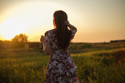 A young girl with long dark hair, in a beautiful dress, looks at the sunset.