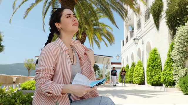 Young Caucasian pretty woman is sitting on bench fanning herself with paper map because of high hot temperature. Summer season. Palms in background. Concept of overheating and sunstroke