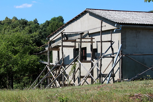 Industrial storage hangar with two small windows and homemade wooden scaffolding prepared for facade reconstruction on side of small hill covered with uncut grass next to dense forest on warm sunny summer day