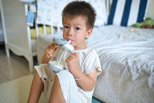 Cute adorable multiracial kid boy sitting on the floor drinking milk from kids bottle. Healthy eating drinking for little children. Supplementary food for growing babies. Candid real authentic moment.