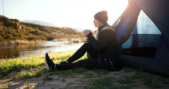 Nature, adventure and woman with coffee on camp sitting and watching the sunrise by her tent. Calm, relax and young female person drinking cappuccino by an outdoor lake in the morning on weekend trip