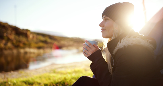 Camping, camper and woman with coffee on adventure sitting and watching the sunrise by her tent. Calm, relax and young female person drinking cappuccino by an outdoor lake in morning on weekend trip.