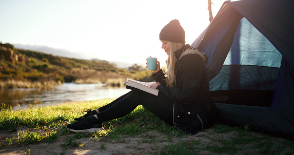 Camping, reading and woman with coffee by tent for adventure, holiday and vacation in morning. Countryside, travel and female person with book at sunrise on mountain, nature and lake for freedom