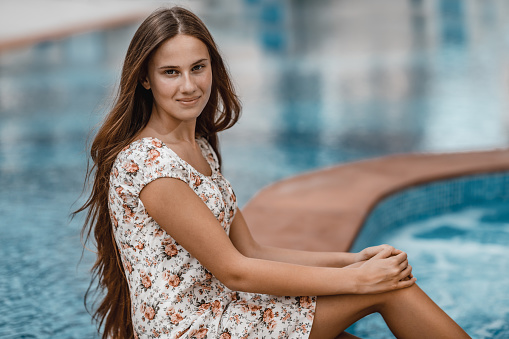 Portrait of a beautiful young female sitting near pool. Spending summer vacation on the beach resort. Happy holidays.
