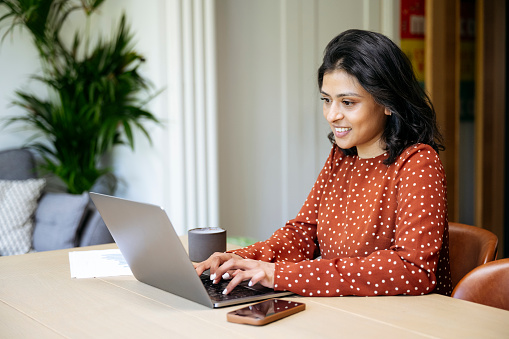 Indian woman in early 40s wearing polka dot dress, sitting at dining table with coffee, printouts, smart phone, and smiling while using laptop.