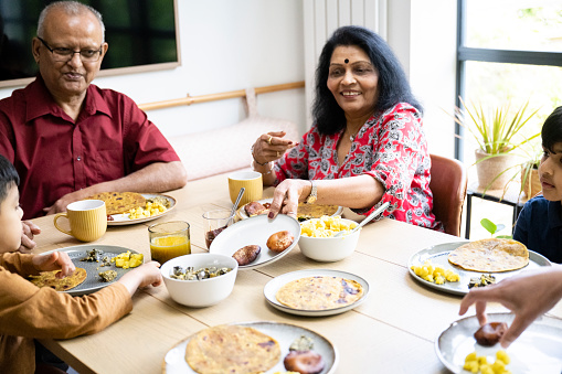Senior Indian couple seated at dining table with 5 and 7 year old boys, smiling, conversing, and eating traditional food.